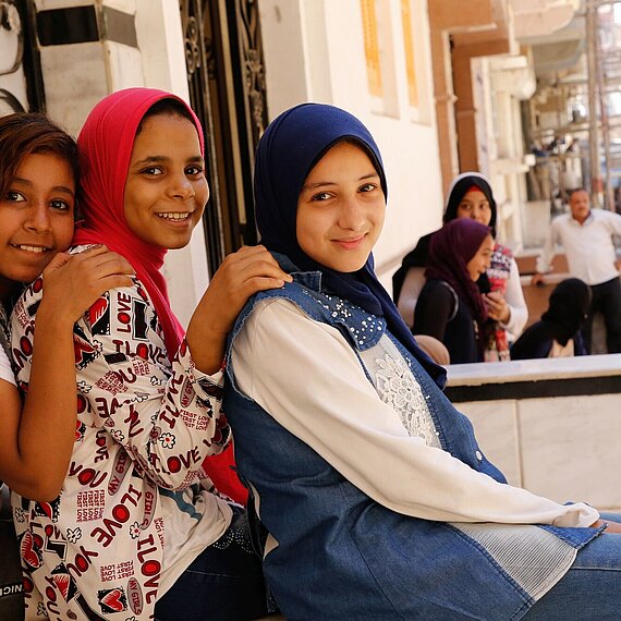 Drei Mädchen lächeln in die Kamera, im Hintergrund weitere Jugendliche auf einer Straße in Ägypten