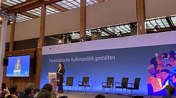 Auf dem Bild steht Ministerin Annalena Baerbock auf der Bühne und spricht zu einem Publikum. Im Hintergrund steht der Schriftzug "Feministische Außenpolitik gestalten"