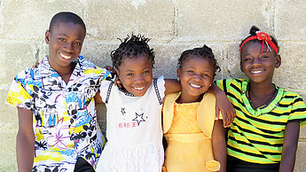 Vier afrikanische Kinder stehen nebeneinander, haben ihre Arme jeweils um die Schultern des neben sich stehenden Kindes gelegt, und lächeln in die Kamera