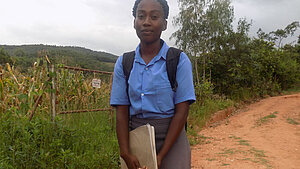 Schülerin mit Schuluniform und Rucksack hält Bücher und steht auf einem ländlichen Weg vor Feldern