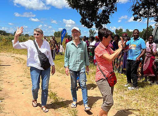 Ehepaar Stackebrandt und eine weitere Mitreisende werden herzlich von einer lokalen Gemeinschaft in Malawi bei einer Projektreise von Plan International begrüßt. Im Hintergrund stehen winkende Menschen und ein Fahrzeug auf sandigem Weg.