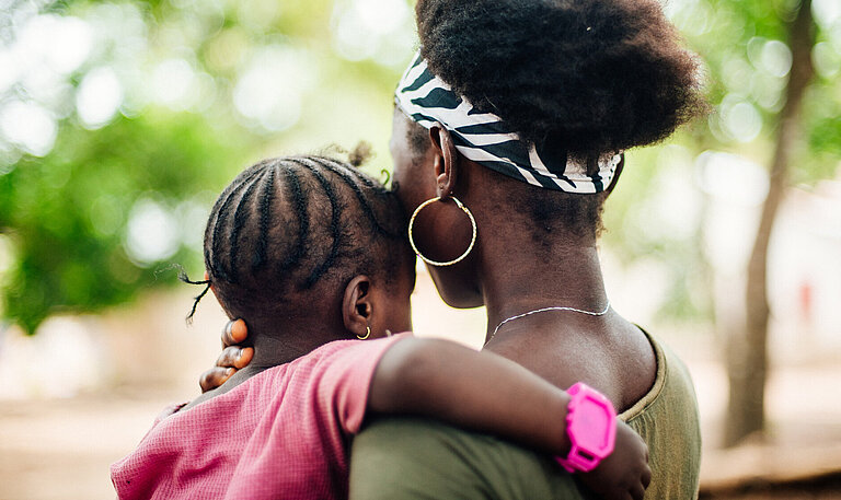Eine junge Mutter mit einem jungen Mädchen auf dem Arm, die beiden sind von hinten zu sehen