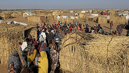 Viele Menschen stehen und gehen zwischen einfachen Hütten aus Stroh und Planen in einer weitläufigen, trockenen Landschaft unter freiem Himmel.