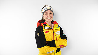 Magdalena Matschine steht vor einem weißen Hintergrund, hat die Arme verschränkt, trägt eine gelbe Skijacke und eine Mütze und lächelt in die Kamera.