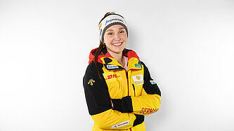 Magdalena Matschine steht vor einem weißen Hintergrund, hat die Arme verschränkt, trägt eine gelbe Skijacke und eine Mütze und lächelt in die Kamera.