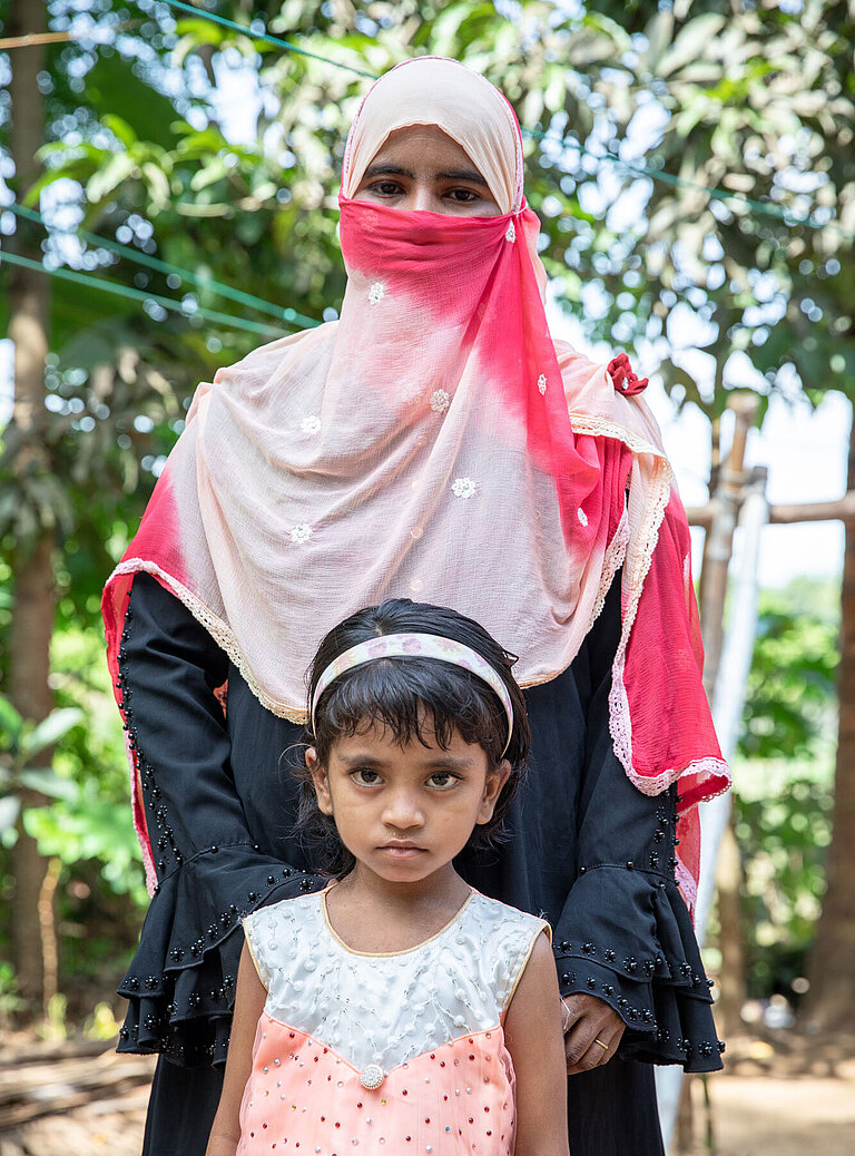 Rozina steht hinter ihrer Tochter Reshmina. Die Mutter trägt eine Nikab und ein Gewand, die Tochter trägt ein rosa Kleid und einen Haarreif. 