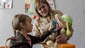 Eine Frau und ein Mädchen spielen mit Puppen in einem Raum, der Teil einer Nothilfeaktion im Kontext des Ukraine-Kriegs ist. An der Wand hängen Kinderbilder.