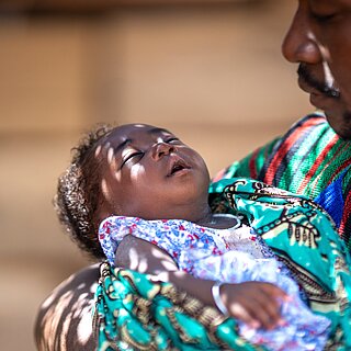 Ein neugeborenes Baby liegt im Arm eines Mannes, der es im Halbschatten in den Schlaf wiegt