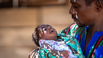 Ein neugeborenes Baby liegt im Arm eines Mannes, der es im Halbschatten in den Schlaf wiegt