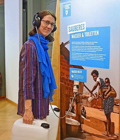 Bettina Gräfin Bernadotte informiert sich in der Ausstellung „Mission 2030“ über das Nachhaltigkeitsziel SDG6 sauberes Wasser Eine Frau in bunter Bluse hebt zwei Kanister mit Wasser