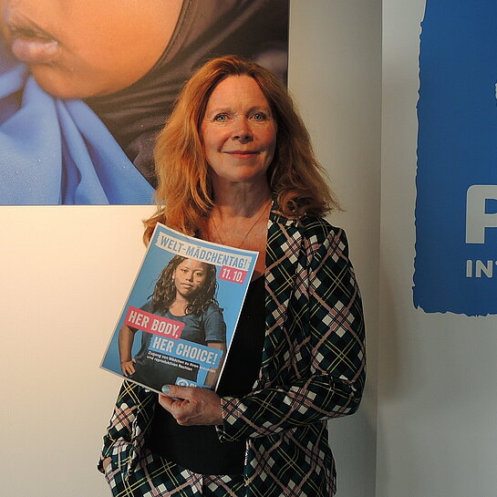 Marion Kracht steht in einem Raum. Im Hintergrund hängt ein großes Bild von einem kleinen Mädchen und das Logo von Plan International