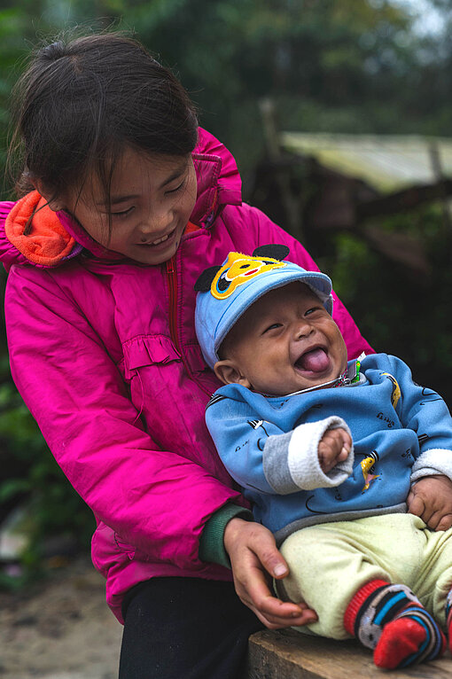 Ein Mädchen spielt mit einem Kleinkind, welches Lacht und die Zunge rausstreckt.