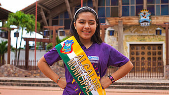 Ein Mädchen trägt eine Schärpe und steht mit den Händen in die Hüften gestemmt vor einem Rathaus in Bolivien