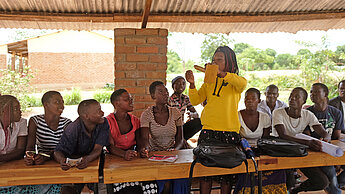 Eine junge Frau steht in Mitten einer Gruppe von Jugendlichen und zeigt ihnen an einem Holz-Modell, wie man ein Kondom benutzt