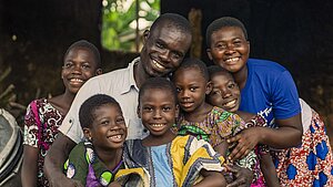 Eine Gruppe von sechs Kindern und einem erwachsenen Mann sitzt eng zusammen und lächelt fröhlich in die Kamera. Die Kinder tragen bunte Kleidung.