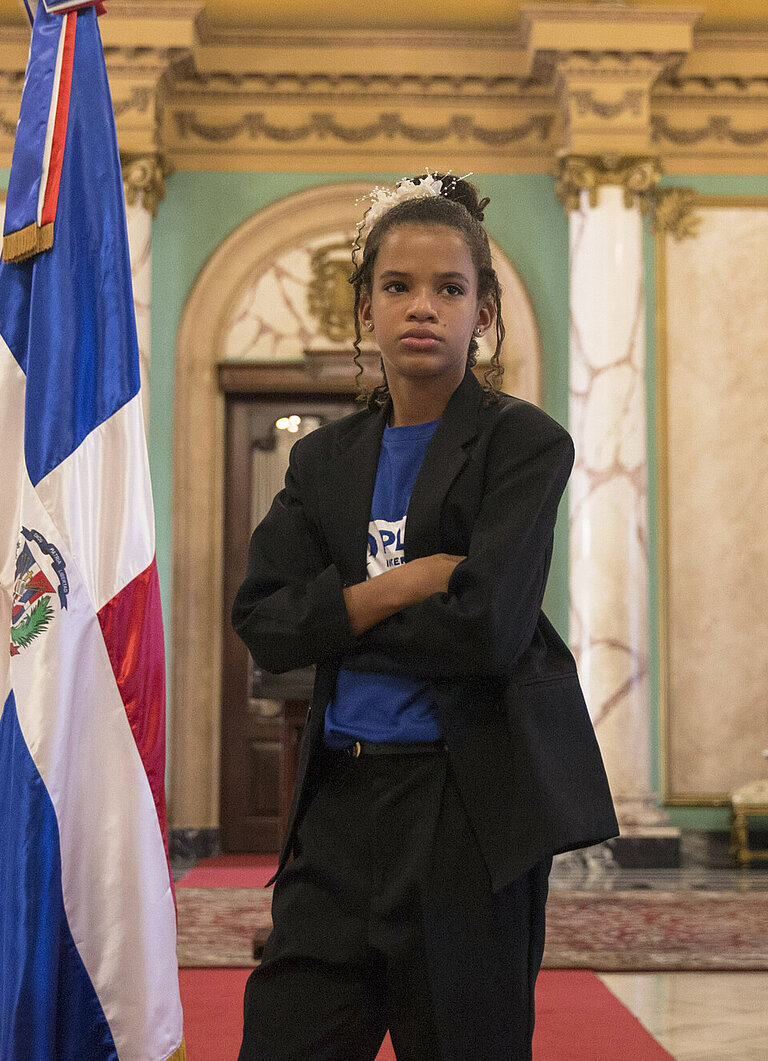 Ein Mädchen steht mit verschränkten Armen in einem Saal, neben ihm die dominikanische Flagge