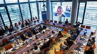 Foto von oben mit Blick auf die Tische, an denen die Teilnehmenden sitzen, und einen großen Bildschirm, auf dem ein Film mit Mädchen aus Simbabwe abgespielt wird