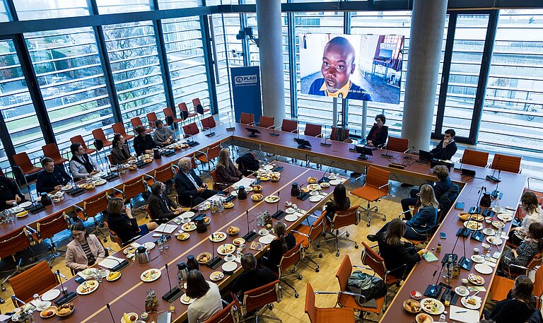 Foto von oben mit Blick auf die Tische, an denen die Teilnehmenden sitzen, und einen großen Bildschirm, auf dem ein Film mit Mädchen aus Simbabwe abgespielt wird