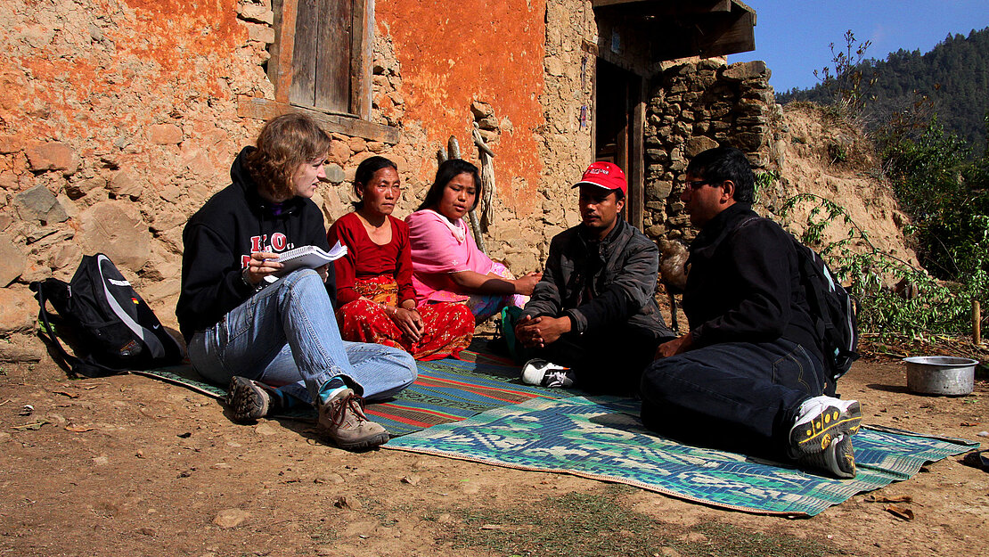 Annika Schönebeck bei Freiwilligeneinsatz sitzt mit Dorfbewohnern und Shreeram K.C. von Plan International Nepal auf einer Matte vor einem traditionellen Haus in Nepal