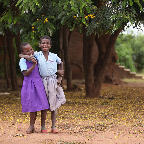 Zwei Schulkinder in Malawi lachen und umarmen sich in einer Umgebung voller Bäume und Blüten. Zwei lachende Schulkinder in Schuluniform umarmen sich unter einem Baum in Malawi und strahlen Freude und Freundschaft aus.