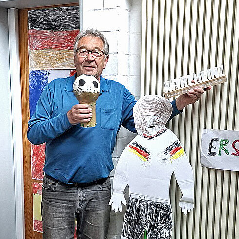 Ein Mann in blauem T-Shirt hält einen World-Cup-Pokal aus Pappe in die Kamera und lacht.
