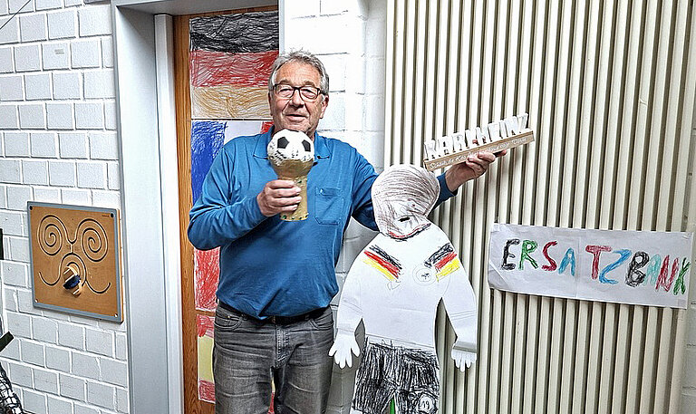 Ein Mann in blauem T-Shirt hält einen World-Cup-Pokal aus Pappe in die Kamera und lacht.