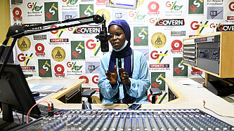 Eine junge Frau sitzt vor einem großen Mischpult und spricht in ein Mikrofon