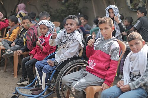 Eine Gruppe von Jungen sitzt im Freien auf Stühlen, einige tragen Schals. Ein Junge sitzt in einem Rollstuhl, mehrere Kinder zeigen mit dem Daumen nach oben.