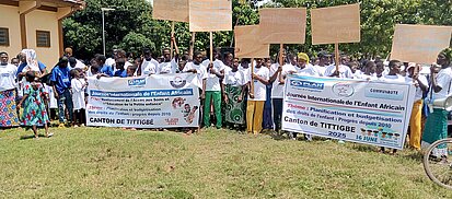 Eine große Gruppe von Kindern und Erwachsenen steht auf einer Wiese vor einem Gebäude. Viele tragen weiße T-Shirts. Mehrere Personen halten Transparente und Schilder mit Text. Die Gruppe ist in Reihen angeordnet, einige Schilder werden hochgehalten.