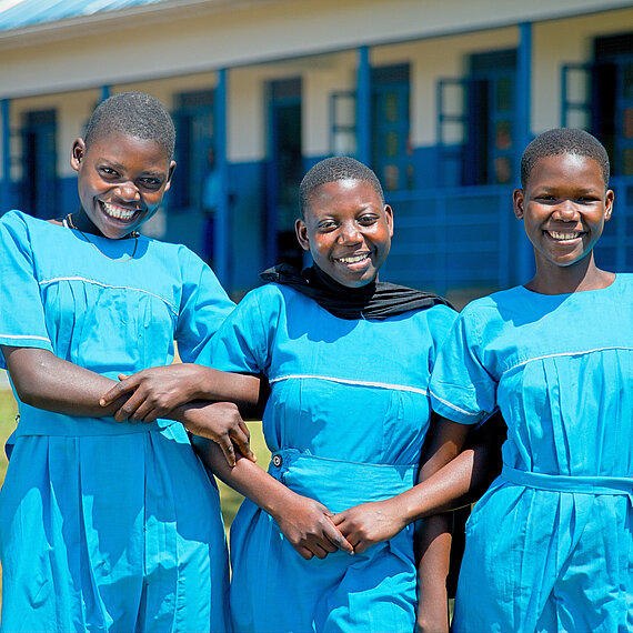 Drei Schülerinnen in blauen Schuluniformen stehen gemeinsam auf dem Schulhof vor einem Schulgebäude