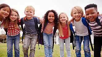  Fröhliche Gruppe von Grundschulkindern mit Rucksäcken, Arm in Arm, im Freien
