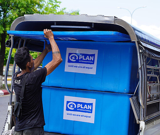 In großen Kisten werden Wasserkanister in Notunterkünfte ausgeliefert Ein Mann verstaut große blaue Boxen voller Wasserkanister, die mit dem Plan-Logo bedruckt sind, in einem Lieferwagen