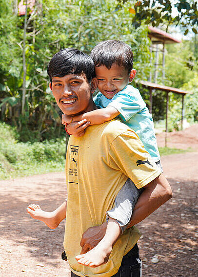 Ein Mann trägt ein junges Kind auf dem Rücken. Beide lächeln. 