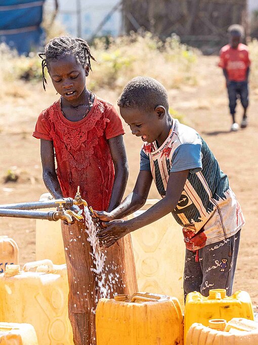 Kinder stehen an einem offenen Wasserhahn und befüllen gelbe Kanister mit Wasser