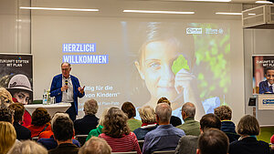 Dr. Werner Bauch, Vorstandsvorsitzender der Stiftung Hilfe mit Plan, spricht bei einer Veranstaltung zum Thema FUTURE-Fonds, Kinderrechte und Klimawandel. Im Hintergrund ist ein großes Bild eines Mädchens zu sehen, das ein Blatt vor seinem Auge hält.