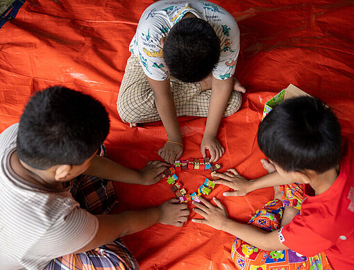 Drei Jungen spielen im Kinderschutzraum in Sagaing Drei Kinder spielen auf einer orangefarbenen Matte ein Spiel zusammen