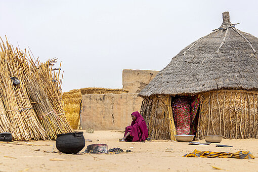 Eine junge Frau in rotem Kleid und Kopftuch sitzt am Eingang einer Hütte. Im Hof stehen noch ein Kessel und große Schüsseln.