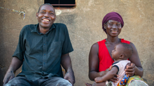  Lächelndes Elternpaar mit Baby auf dem Schoß vor einem Haus in Westafrika – Szene positiver Elternschaft.