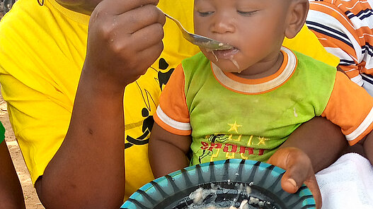 Eine Frau füttert das kleine Kind, das auf ihrem Schoß sitzt.