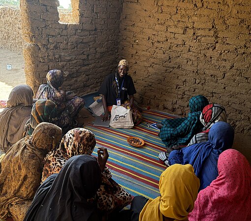 Eine Mitarbeiterin von Plan International sitzt mit mehreren Frauen im Gespräch vor einer Unterkunft aus Stroh.