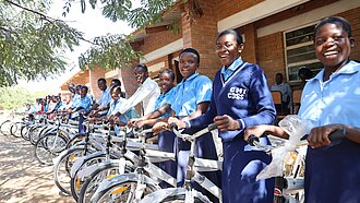Eine Gruppe von Schülerinnen und Schülern, die lächelnd neben neu verteilten Fahrrädern vor einem Schulgebäude in Malawi stehen. Die Schülerinnen und Schüler tragen Schuluniformen und die Fahrräder sind bereit zur Nutzung.