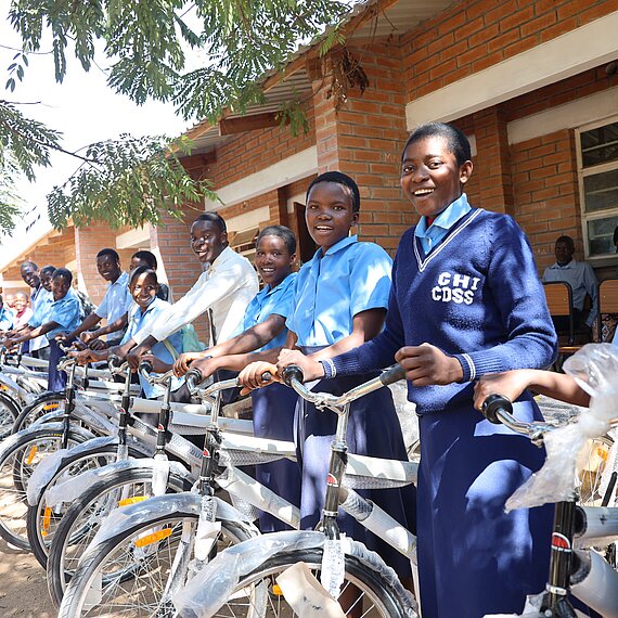 Eine Gruppe von Schülerinnen und Schülern, die lächelnd neben neu verteilten Fahrrädern vor einem Schulgebäude in Malawi stehen. Die Schülerinnen und Schüler tragen Schuluniformen und die Fahrräder sind bereit zur Nutzung.