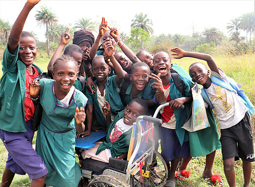 Schulkinder Sierra Leone Eine Gruppe Schulkinder schauen in die Kamera, lachen und heben die Arme. In der Mitte steht ein Junge mit Rollstuhl, der auch in die Kamera lacht.
