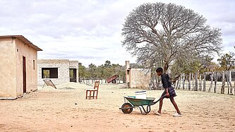 Kimberly schiebt eine Schubkarre über Sand, darauf steht ein großer Eimer
