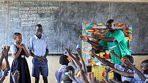 Lehrer leitet eine interaktive, spielerische Unterrichtsstunde mit Kindern in einer Schule in einem Flüchtlingslager in Uganda. Die Schülerinnen und Schüler beteiligen sich aktiv und arbeiten mit bunten Bausteinen zur Förderung des kindzentrierten Lernens.