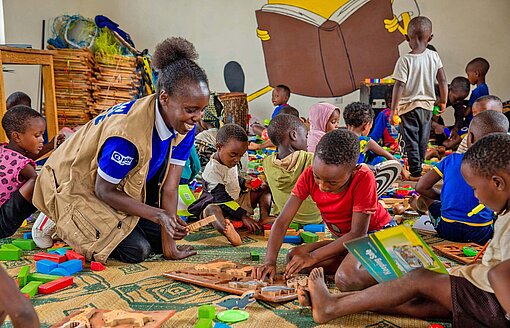 Eine Frau sitzt lächelnd inmitten von kleinen Kinder in einem Zimmer und spielt mit ihnen