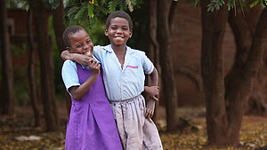 Zwei lachende Schulkinder in Schuluniform umarmen sich unter einem Baum in Malawi und strahlen Freude und Freundschaft aus.