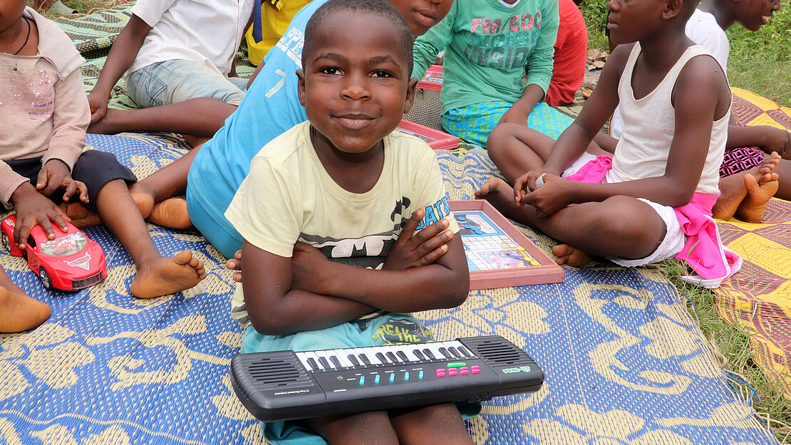 Ein kleiner Junge sitzt gemeinsam mit anderen Kindern auf einer Matte auf dem Boden. Auf dem Schoß hat er ein kleines Keyboard.