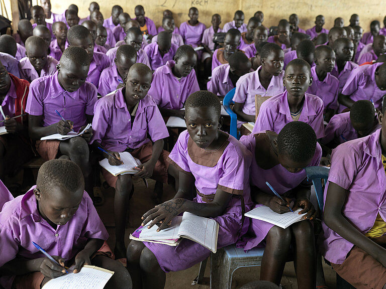 Schüler:innen mit lilafarbener Uniform sitzen im Klassenzimmer und schreiben fleißig mit.