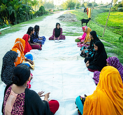 Eine junge Frau leitet Gespräch mit Mädchen einer Jugendgruppe in Bangladesch; sie sitzen für einen Workshop zum Thema Kinderehe und Mädchenrechte im Kreis auf einer Decke im Freien.